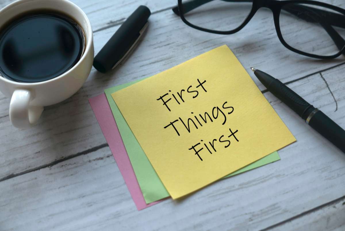 Selective focus of a cup of coffee, pen, glasses and memo notes written with First Things First on white wooden background