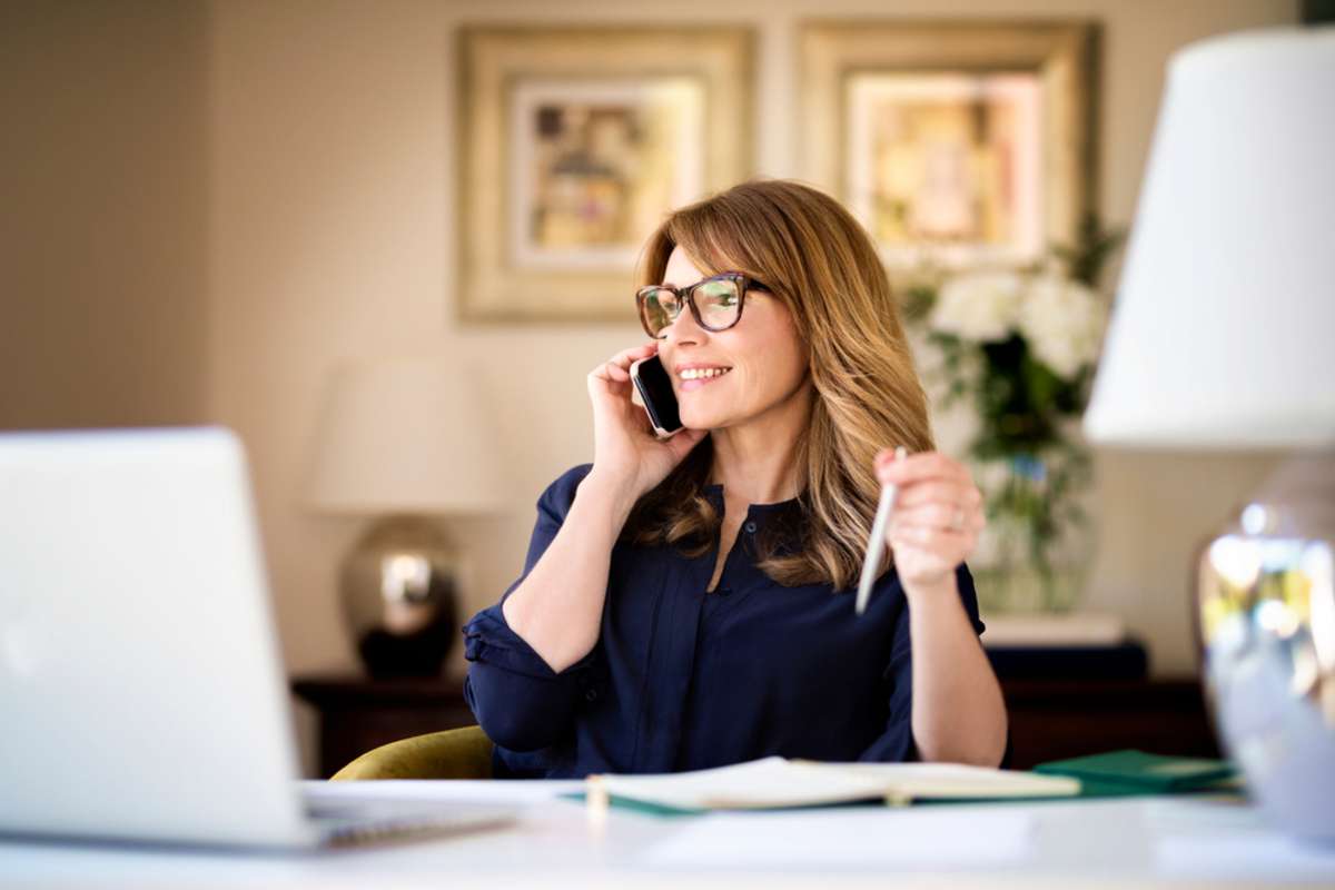 Shot happy businesswoman sitting at desk behind her laptop and talking with somebody on her mobile phone while working from home