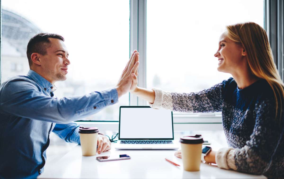 Successful young male and female colleagues giving five each other for great collaborative job during meeting at table